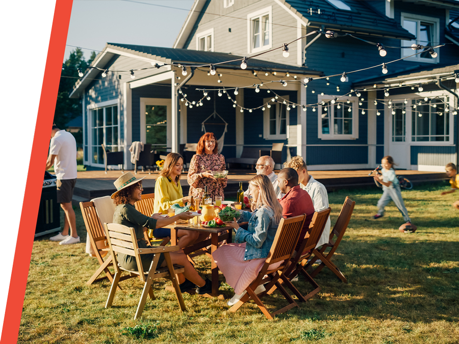 Family and friends enjoying a backyard dinner under string lights on a sunny evening beside a modern house.