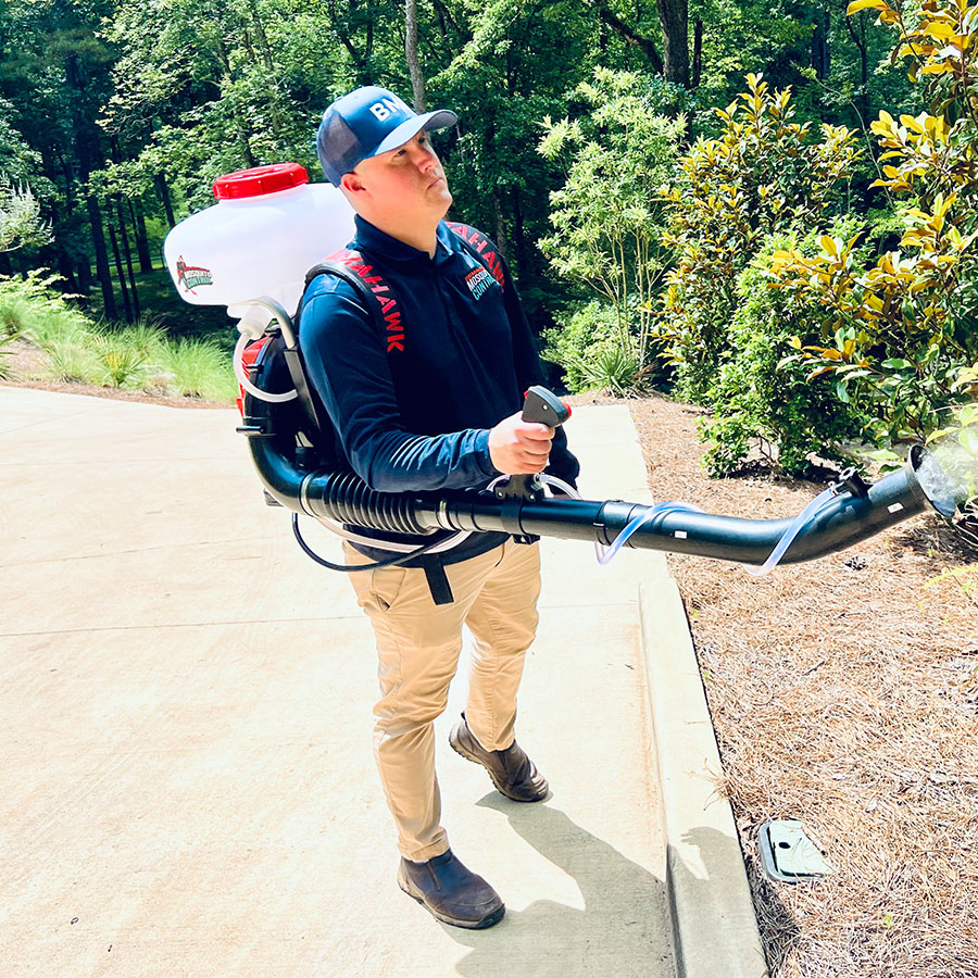 A man landscaper wearing a cap using a backpack blower to clear driveway edge and mulch bed near shrubs